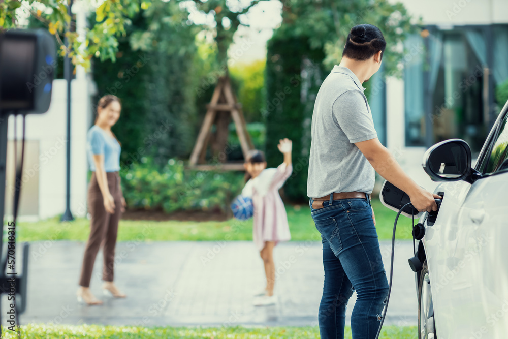 Focus image of progressive man charging electric car from home charging station with blur mother and