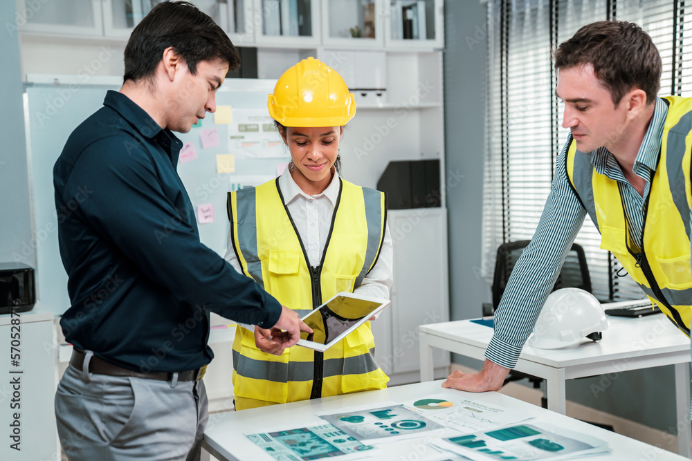 A team of competent engineers wearing safety equipment is working on blueprints with a tablet while 