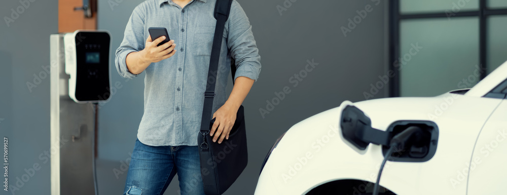 Progressive asian man and electric car with home charging station. Concept of the use of electric ve