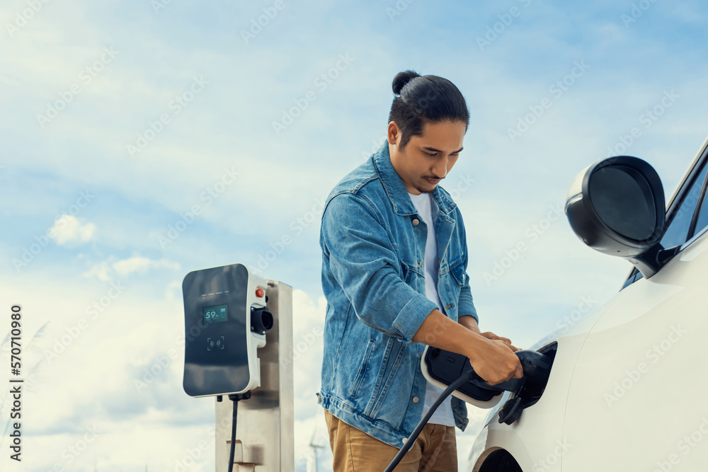 Progressive man with his electric car, EV car recharging energy from charging station on green field