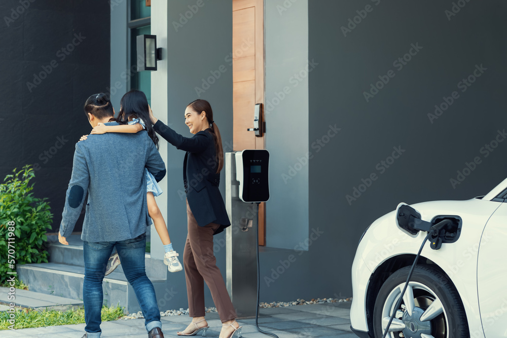 Progressive young parents and daughter with electric vehicle and home charging station. Green and cl