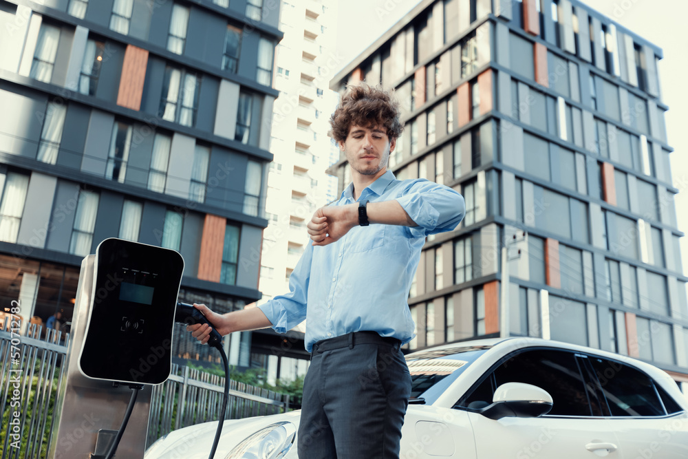 Businessman with smartwatch at modern charging station for electric vehicle with background of resid