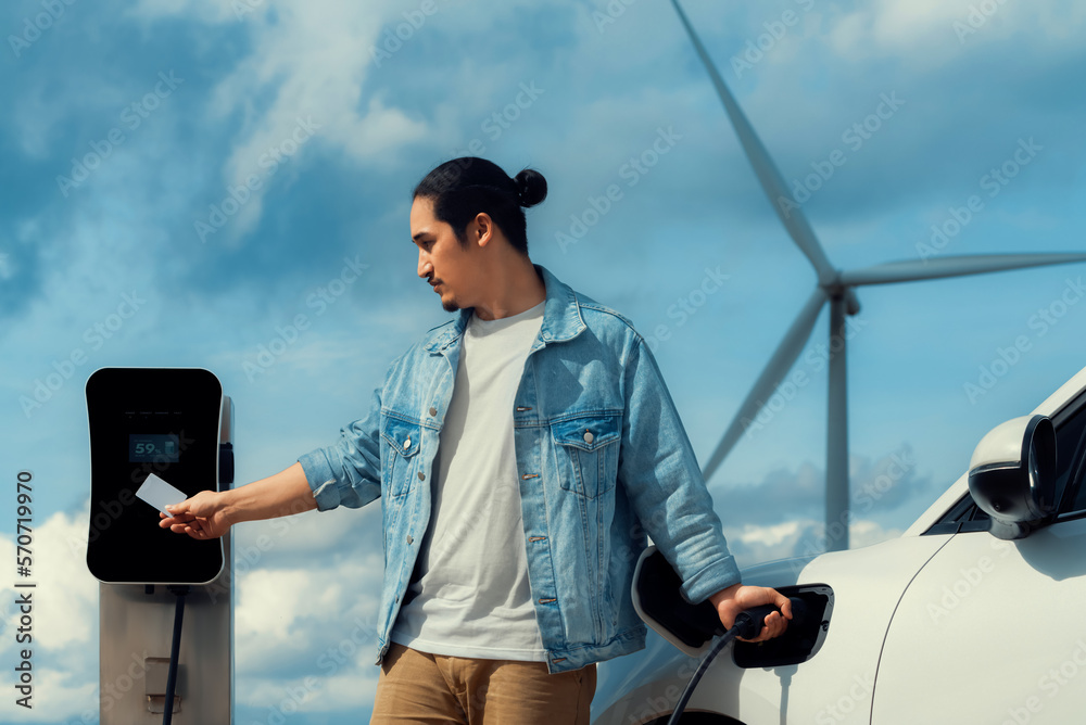 Progressive man with his electric car, EV car recharging energy from charging station on green field