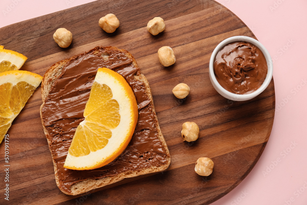 Wooden board of tasty toast with hazelnut butter and orange slice on pink background, closeup