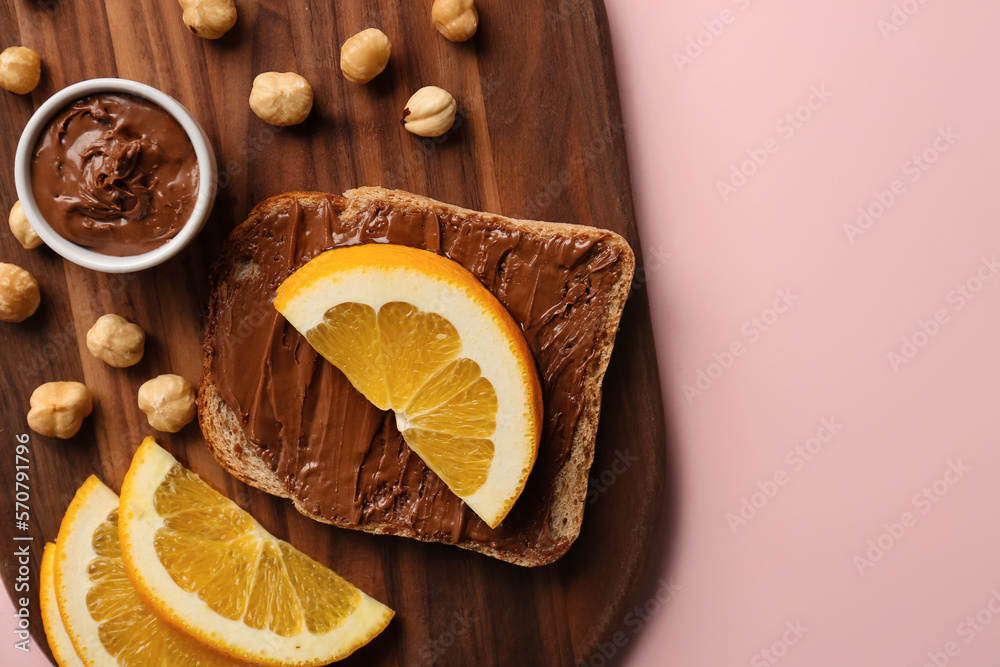 Wooden board of tasty toast with hazelnut butter and orange slices on pink background, closeup