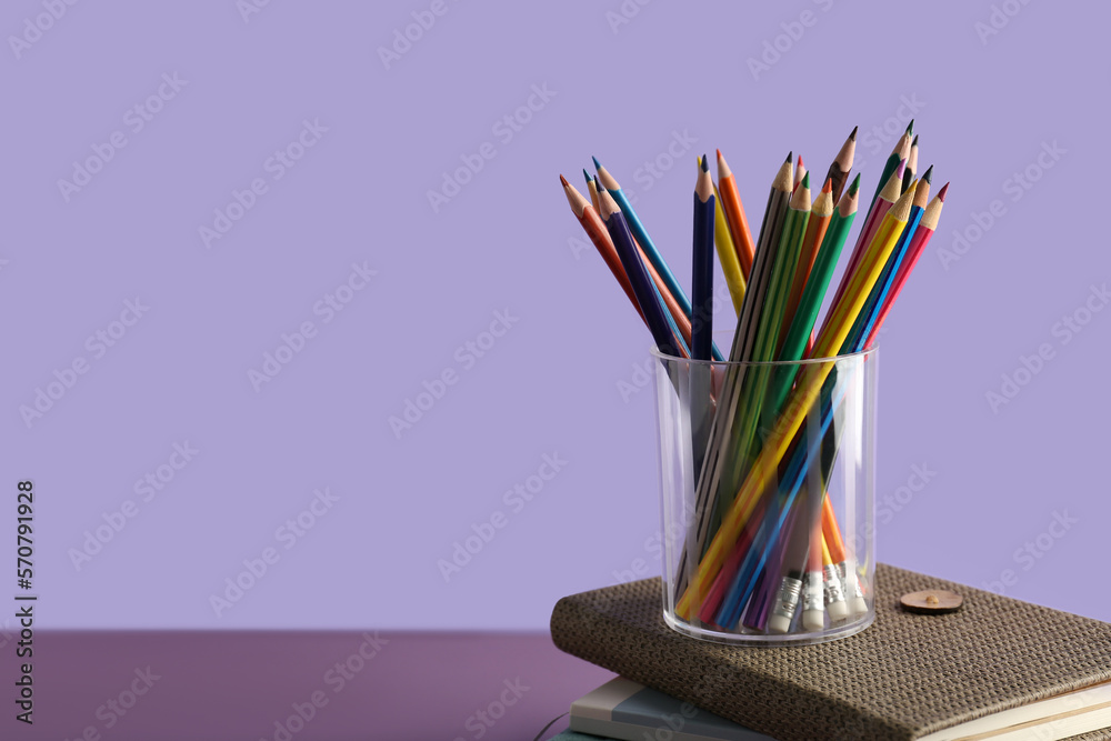 Glass with colorful pencils on stack of notebooks against color background