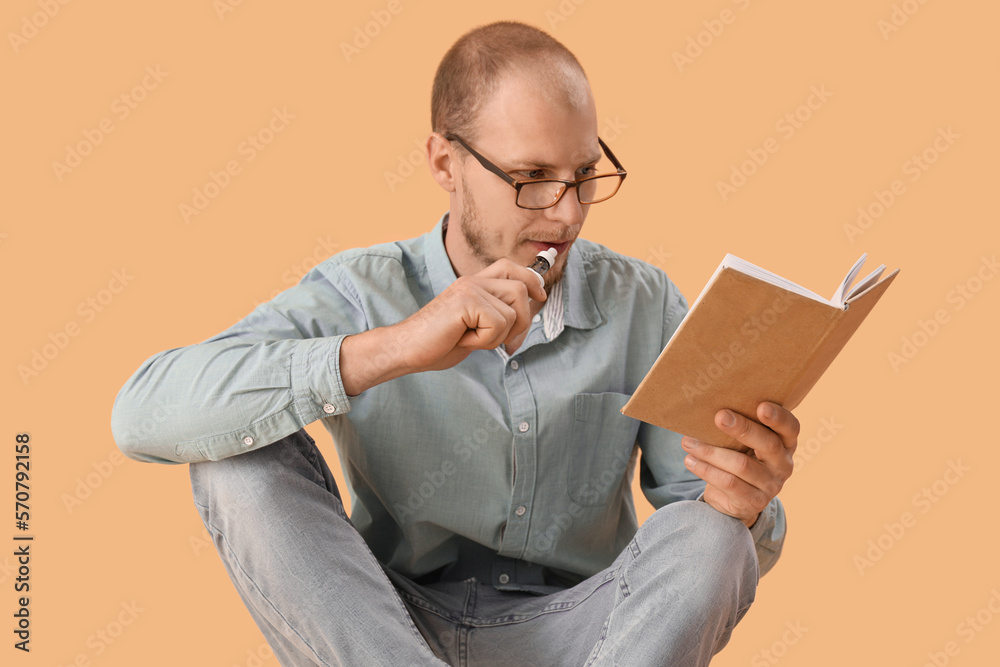 Young man in eyeglasses with disposable electronic cigarette reading book on beige background