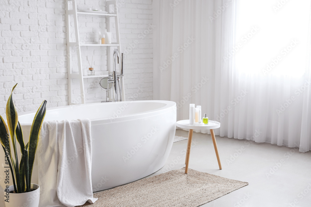 Interior of light bathroom with bathtub, table and shelving unit