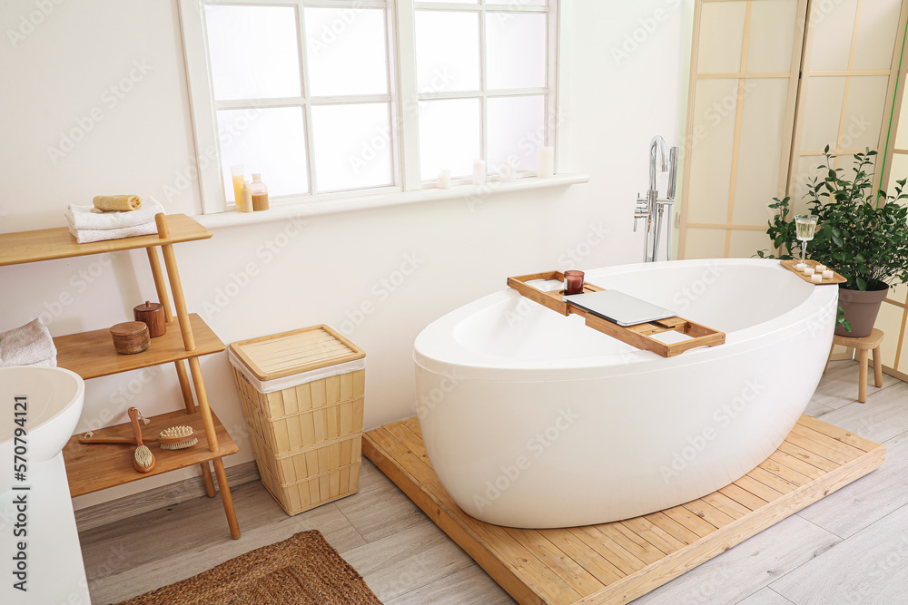 Interior of bathroom with modern bathtub, laptop, champagne and burning candles on board