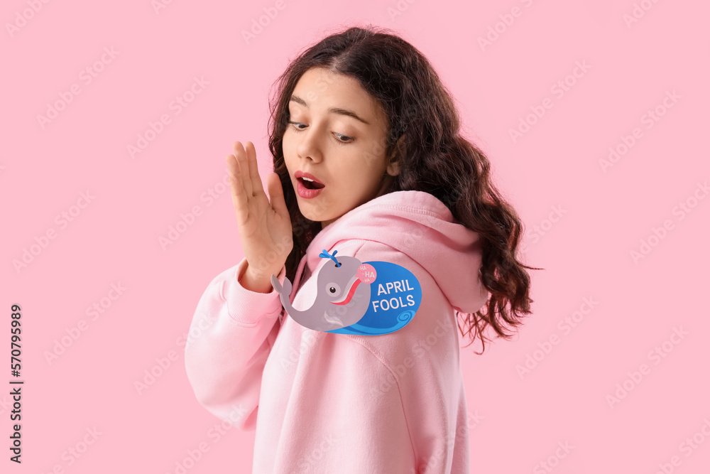 Shocked teenage girl and paper with text APRIL FOOLS on her shoulder against pink background