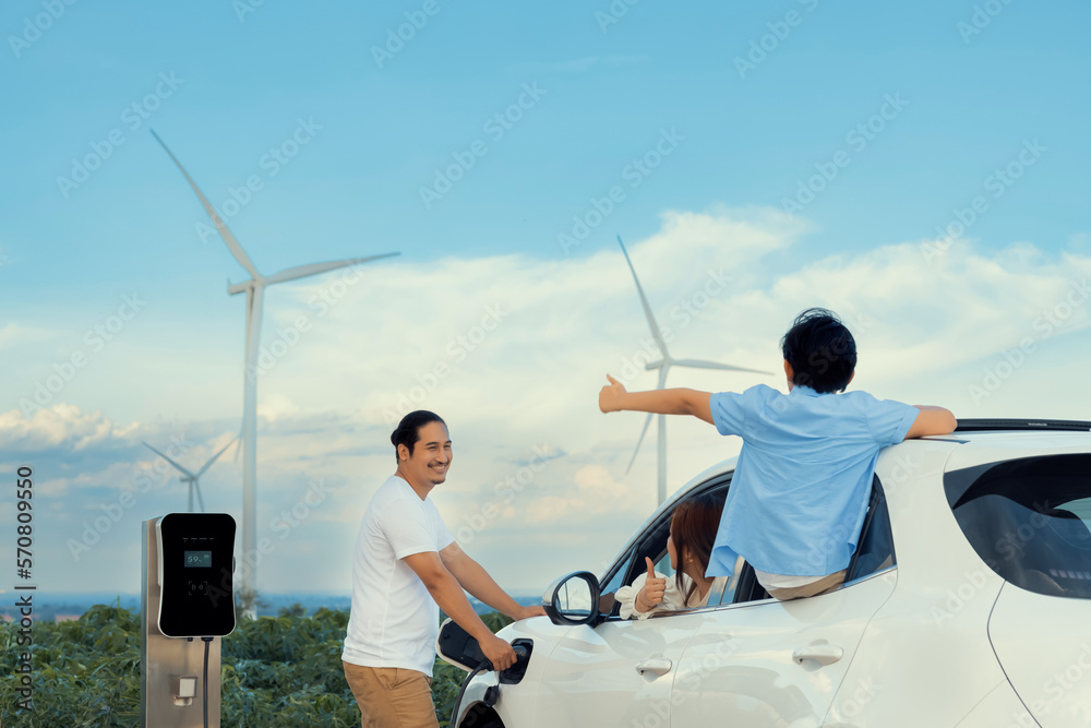 Concept of progressive happy family enjoying their time at wind farm with electric vehicle. Electric