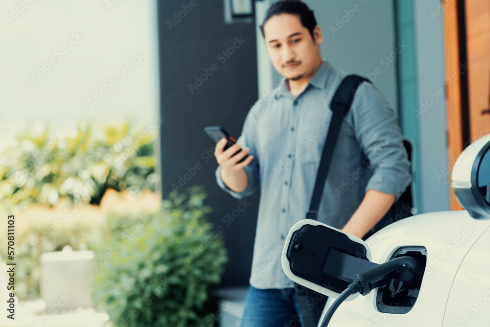 Focus electric car charging at home charging station with blurred progressive man walking in the bac