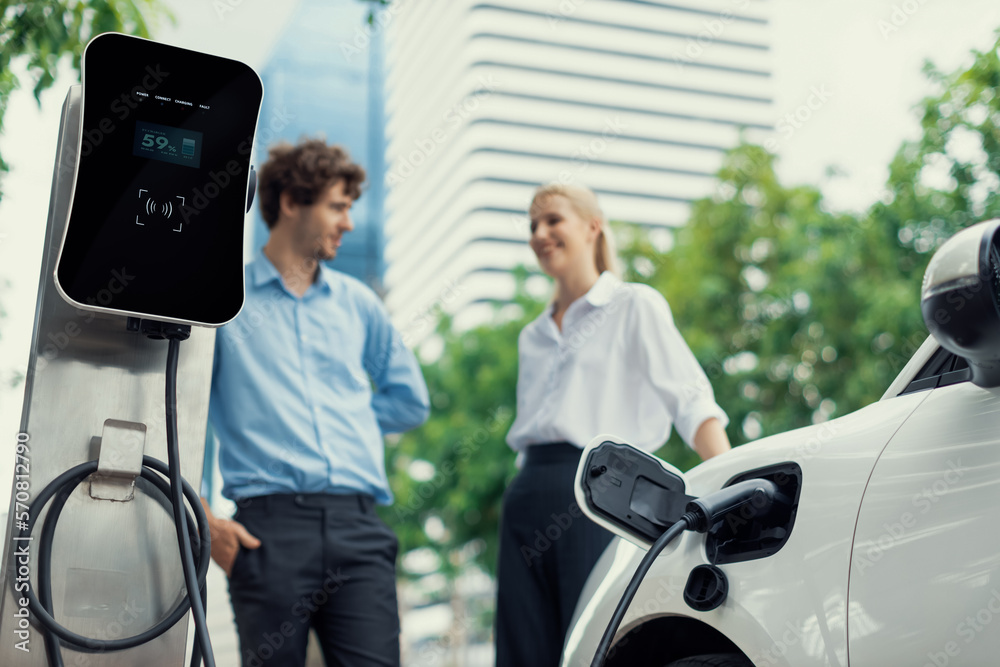 Focus parking-electric car connected to public charging station with blur progressive businesspeople