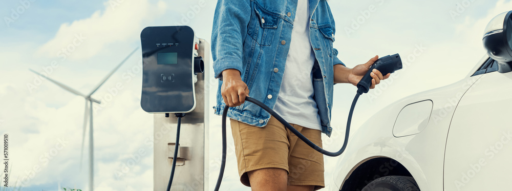 Progressive man with his electric car, EV car recharging energy from charging station on green field