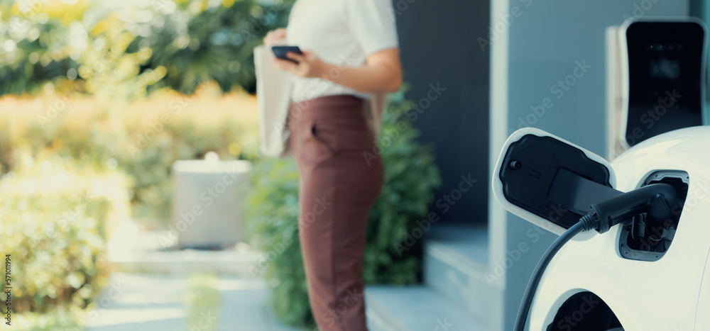 Focus image of electric vehicle recharging battery at home charging station with blurred woman walki