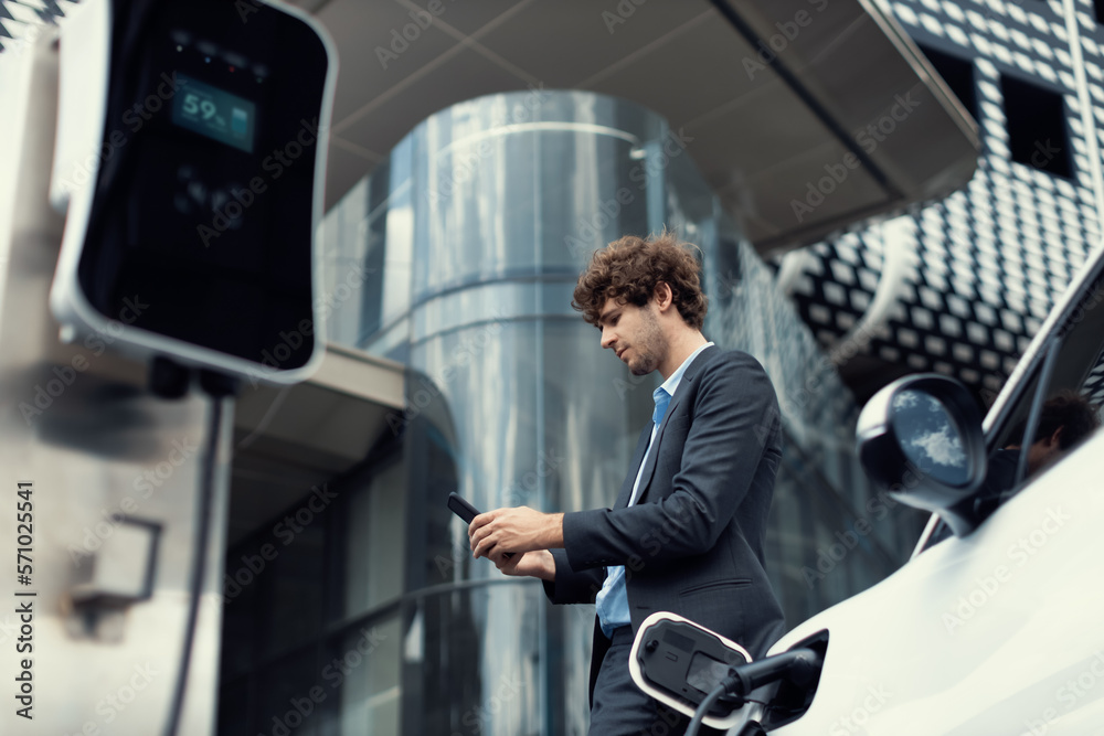 Below view of progressive businessman with electric car recharging at public charging station at mod