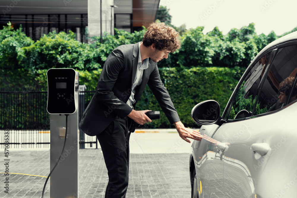 Progressive businessman with electric car recharging at public charging station at modern city resid