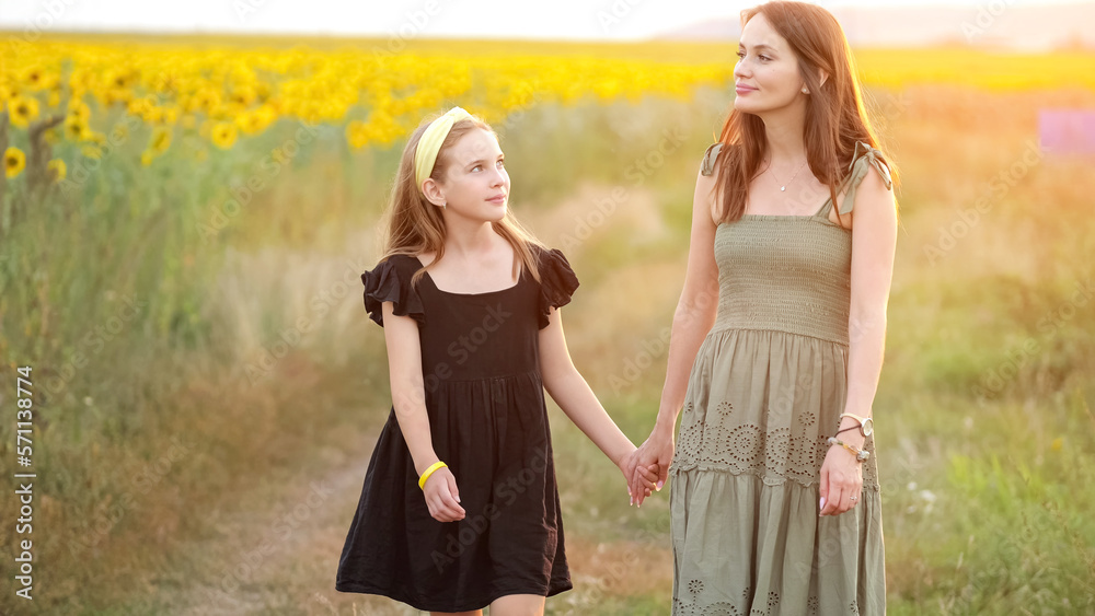 Woman and teenager girl walk together past blooming sunflower field talking about countryside holida