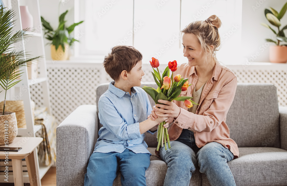 Cute boy sitting on the sofa with mom and giving a bouquet of tulips to her.