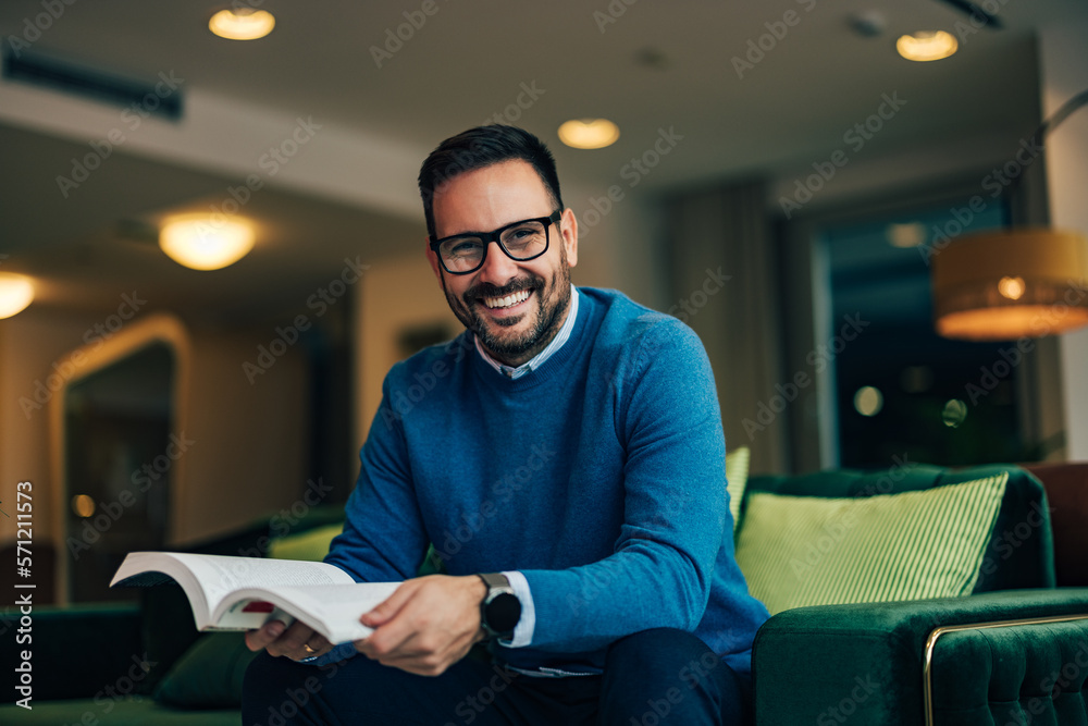 Portrait of a smiling businessman, wearing a blue sweater, sitting on a green couch, and reading a b