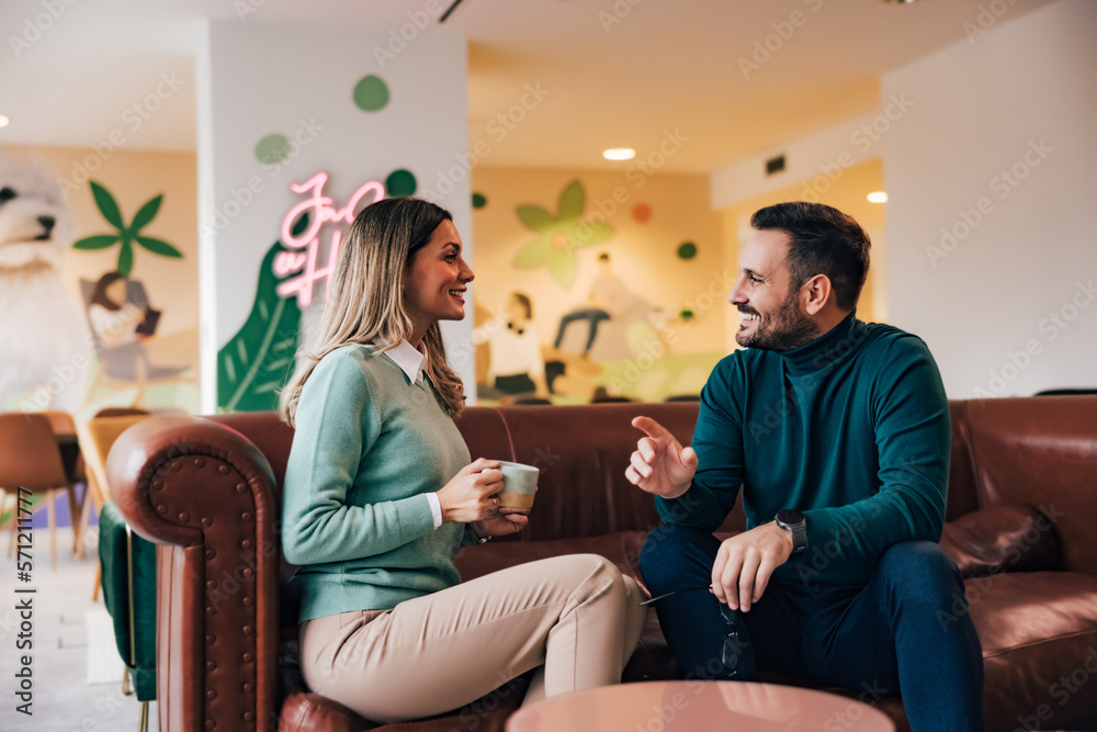 Businesswoman and businessman talking, drinking coffee, smiling, sitting on the leather sofa at the 