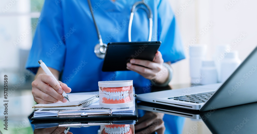 Dentist sitting at table with jaw samples tooth model and working with tablet and laptop in dental o