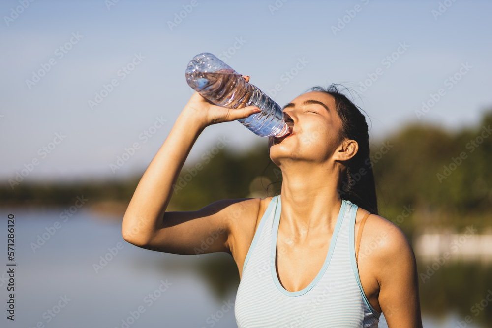 Thirsty Asian female athlete drinking water after exercising outdoors in park.