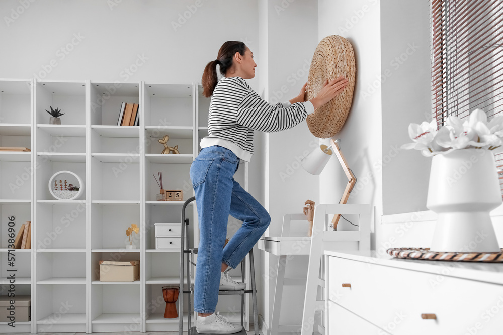 Young woman on stepladder hanging rattan pouf onto wall at home