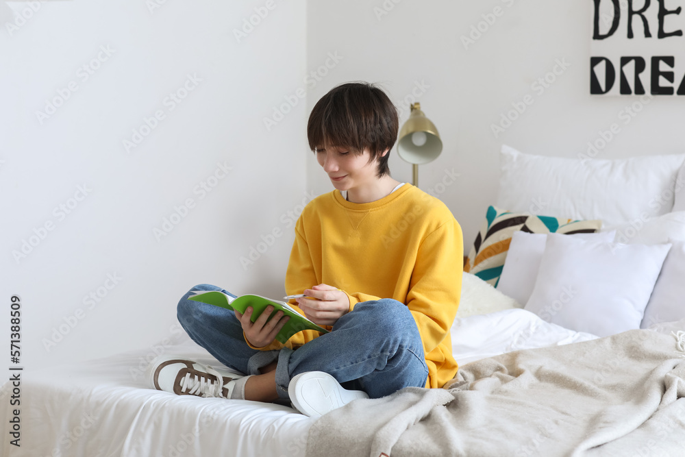 Male student writing in notebook on bed at home