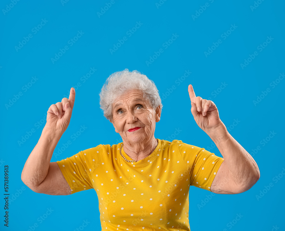 Senior woman in yellow t-shirt pointing at something on blue background