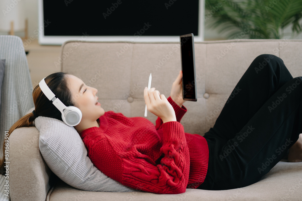 ￼Smiling girl relaxing at home, she is playing music using smartphone tablet, laptop, and wearing wh