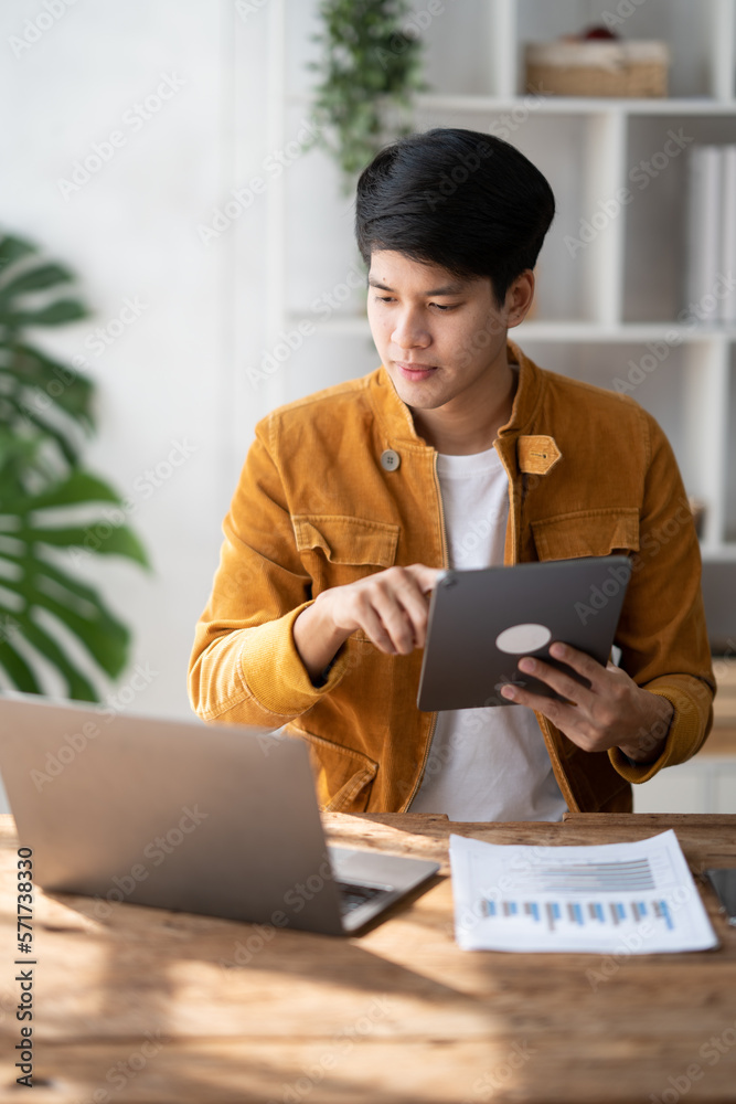 Smiling Asian man working on laptop computer while holding digital tablet in modern office space. .