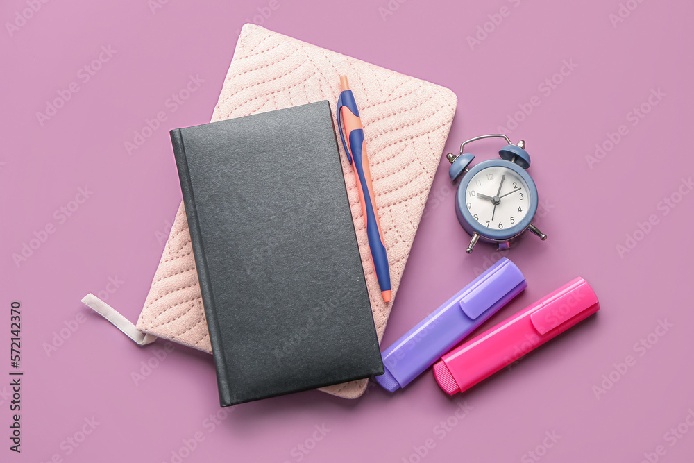 Notebooks, pen, markers and alarm clock on lilac background