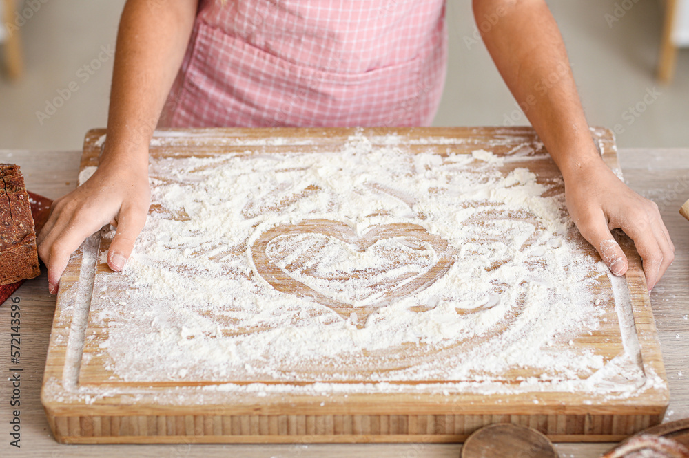 Little baker and board with heart made of flour on table in kitchen, closeup