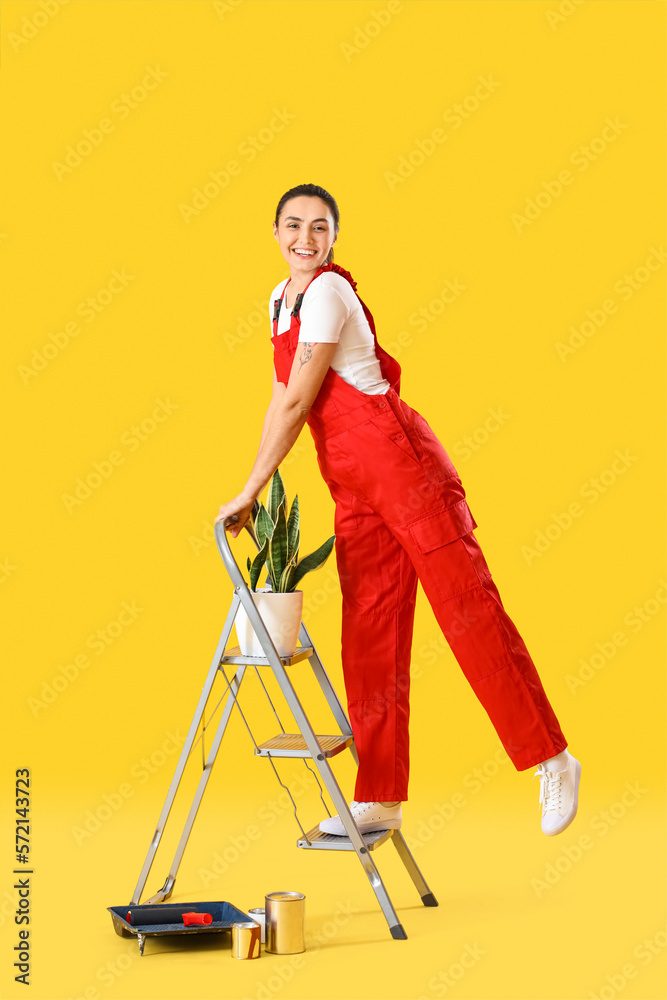 Young woman with houseplant, ladder and paint cans on yellow background