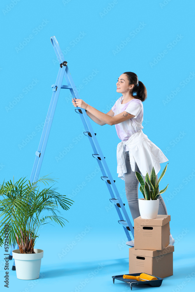 Young woman with ladder and moving boxes on blue background