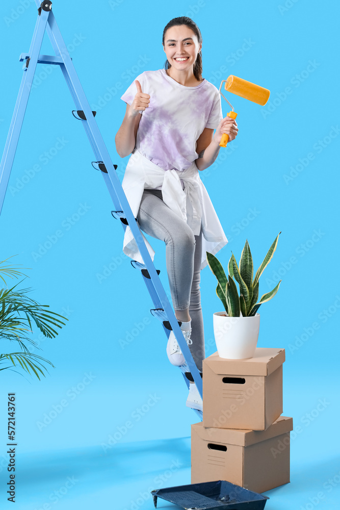 Young woman with paint roller, ladder and moving boxes showing thumb-up on blue background