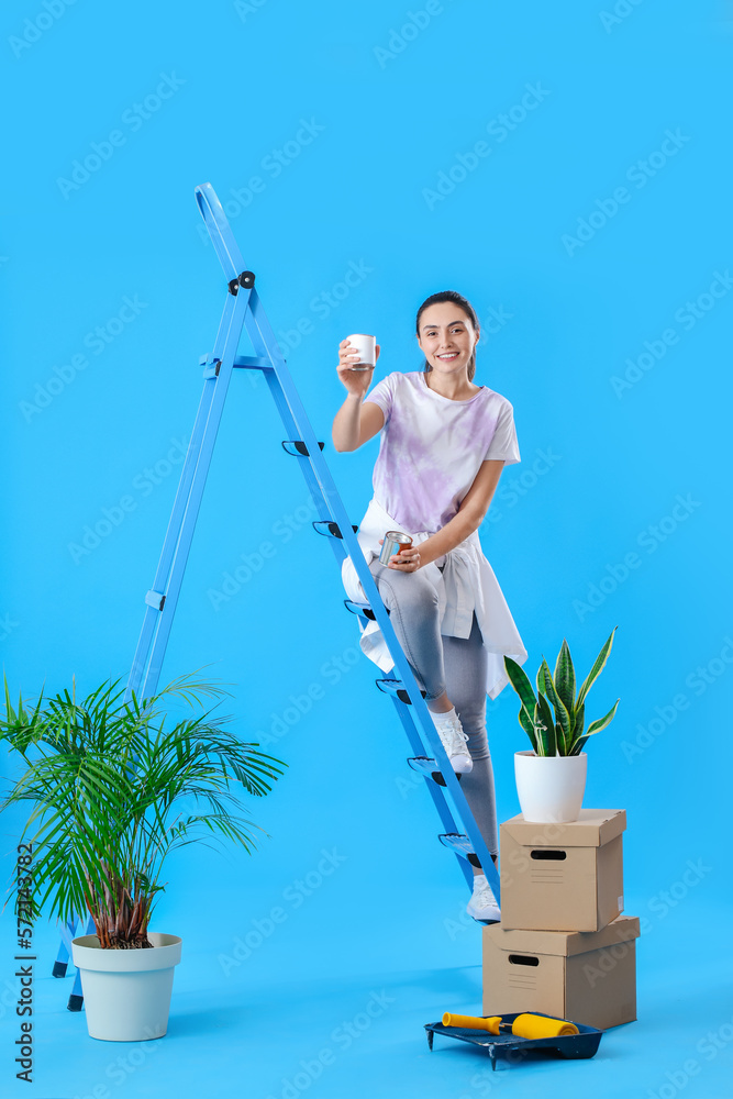 Young woman with paint cans, ladder and moving boxes on blue background