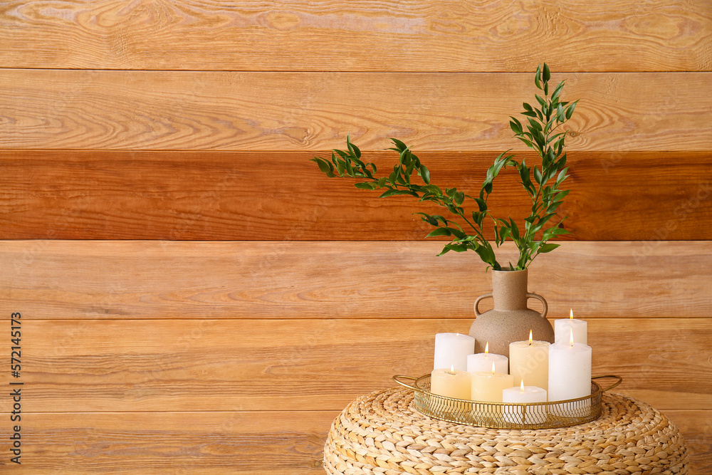 Tray with burning candles and plant branches in vase on pouf near wooden wall