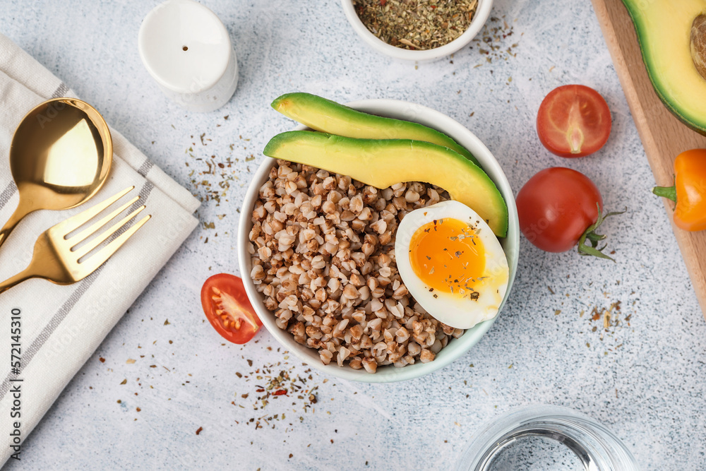 Bowl of tasty buckwheat porridge with soft boiled egg and fresh vegetables on grey table