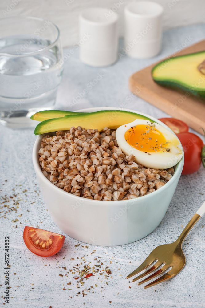 Bowl of tasty buckwheat porridge with soft boiled egg and fresh vegetables on grey table