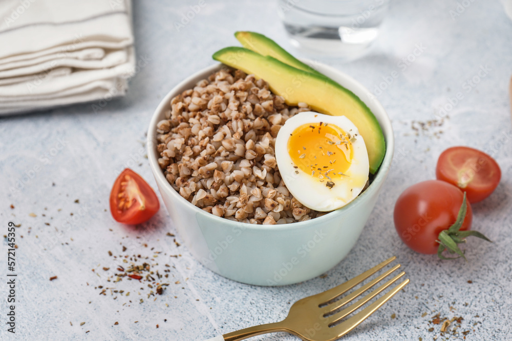 Bowl of tasty buckwheat porridge with soft boiled egg and fresh vegetables on grey table