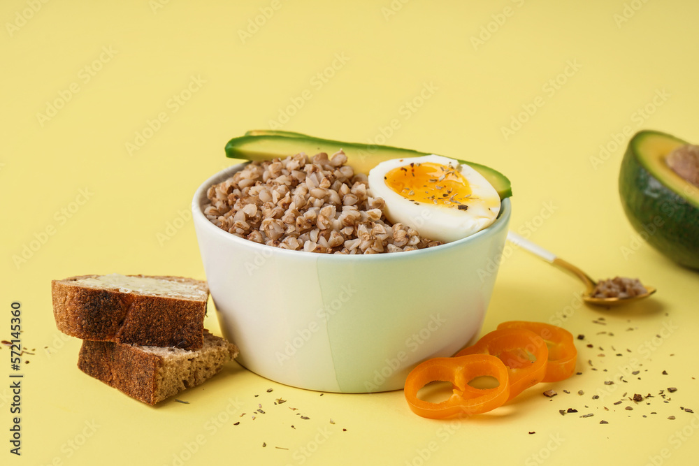 Bowl of tasty buckwheat porridge with soft boiled egg, fresh vegetables and bread on yellow backgrou