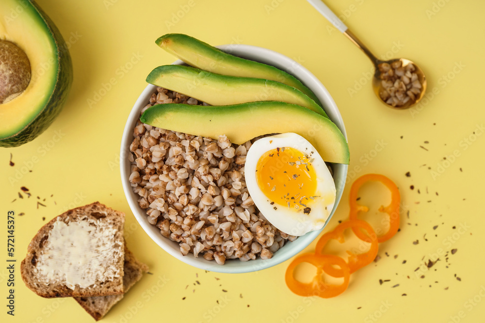 Bowl of tasty buckwheat porridge with soft boiled egg, fresh vegetables and bread on yellow backgrou