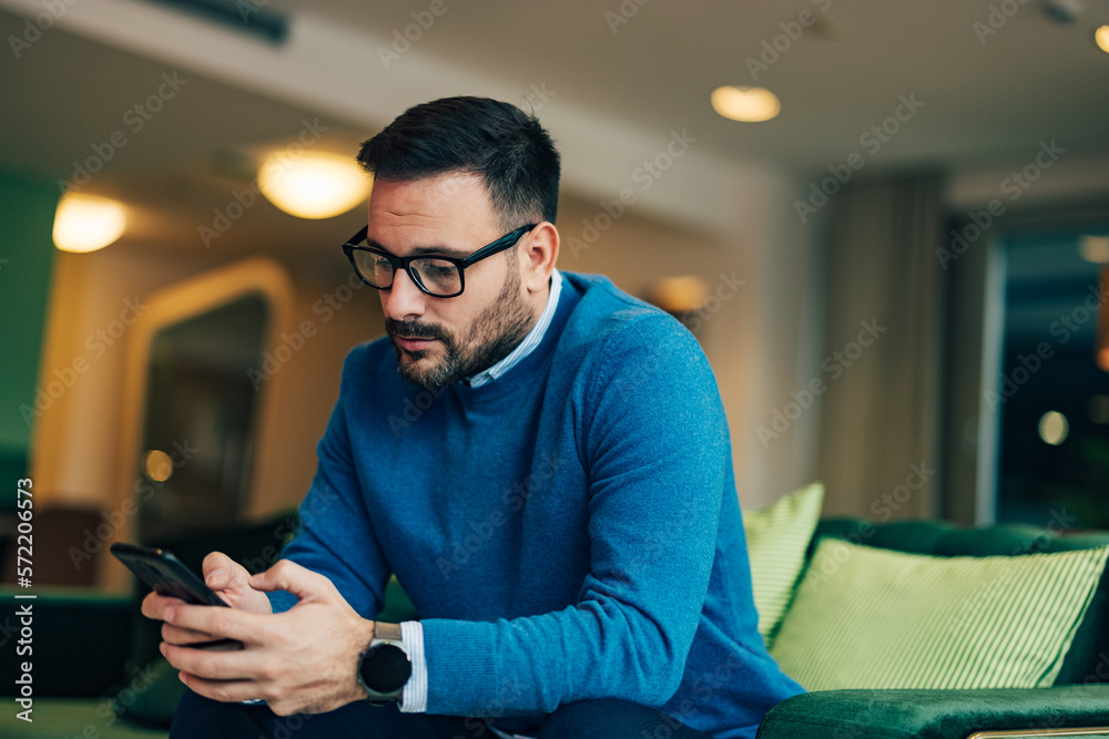 Focused businessman with glasses typing on the phone, wearing a blue sweater, and sitting on the cou