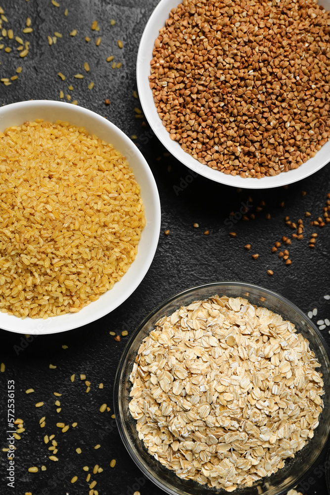 Bowls of buckwheat, bulgur and oatmeal on dark background, closeup