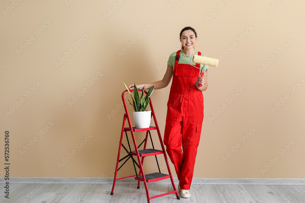 Young woman with paint roller, ladder and houseplant near beige wall