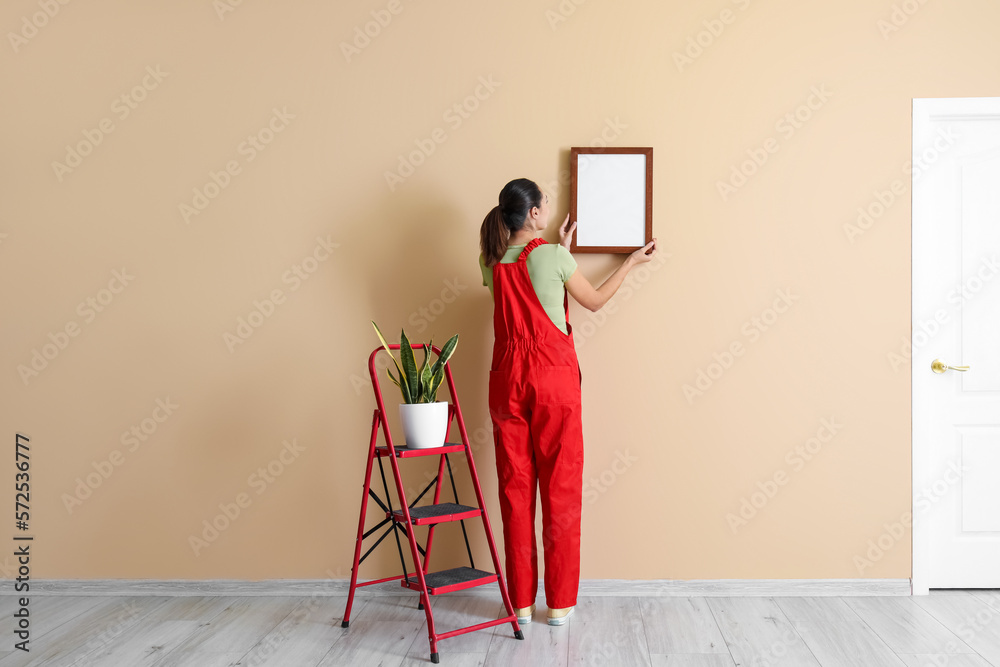 Young woman hanging frame on beige wall, back view