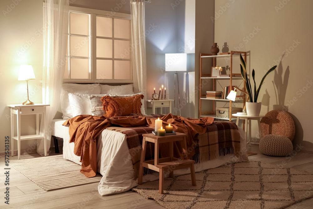 Interior of bedroom with brown checkered blankets on bed, burning candles and glowing lamps late in 