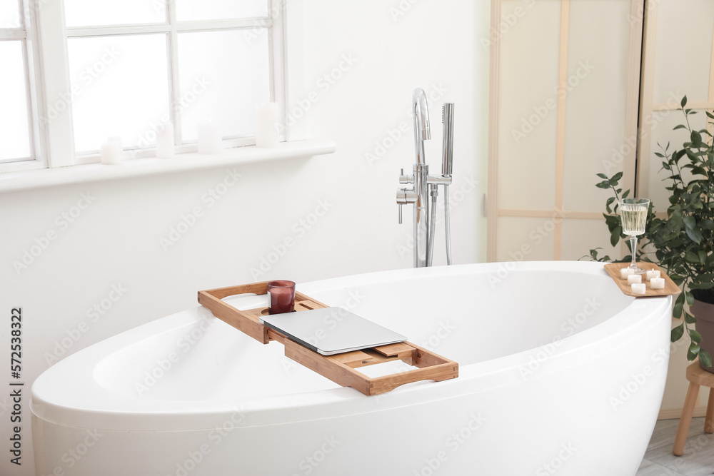 Interior of bathroom with modern bathtub, laptop, champagne and burning candles on board